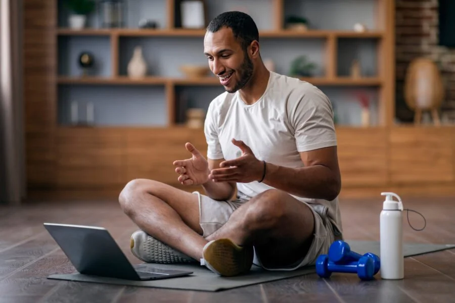 A male professional athlete, sitting cross-legged on a yoga mat in a living room, smiling and engaging with a laptop. Exercise equipment, including blue dumbbells and a water bottle, are placed nearby as he speaks to his branding coach and strategist