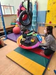 A woman with a mask sitting on the floor in a therapy or playroom for children, engaging with a young child in a swing or therapy seat surrounded by toys, balls, and gym equipment.