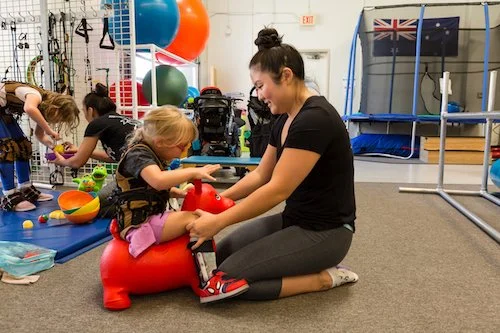 A young girl in a wheelchair playing with a toy while an adult woman kneeling on the floor interacts with her at a therapy or activity center.