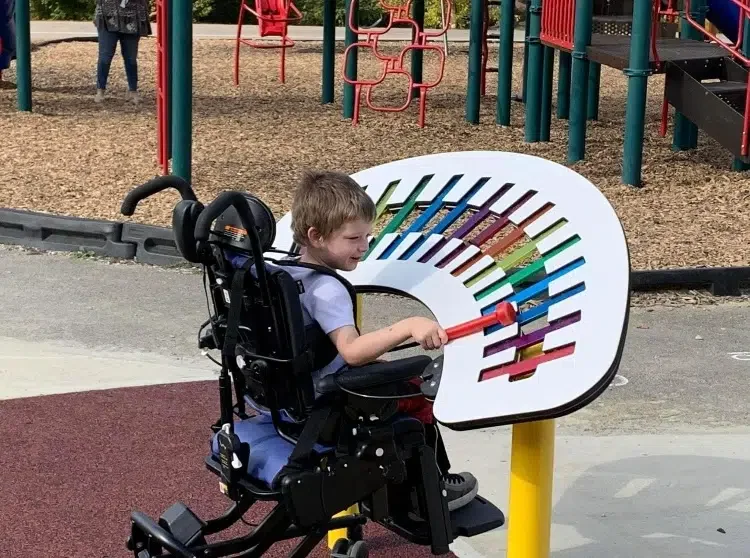 Young boy in a wheelchair playing a rainbow-colored xylophone at a playground.