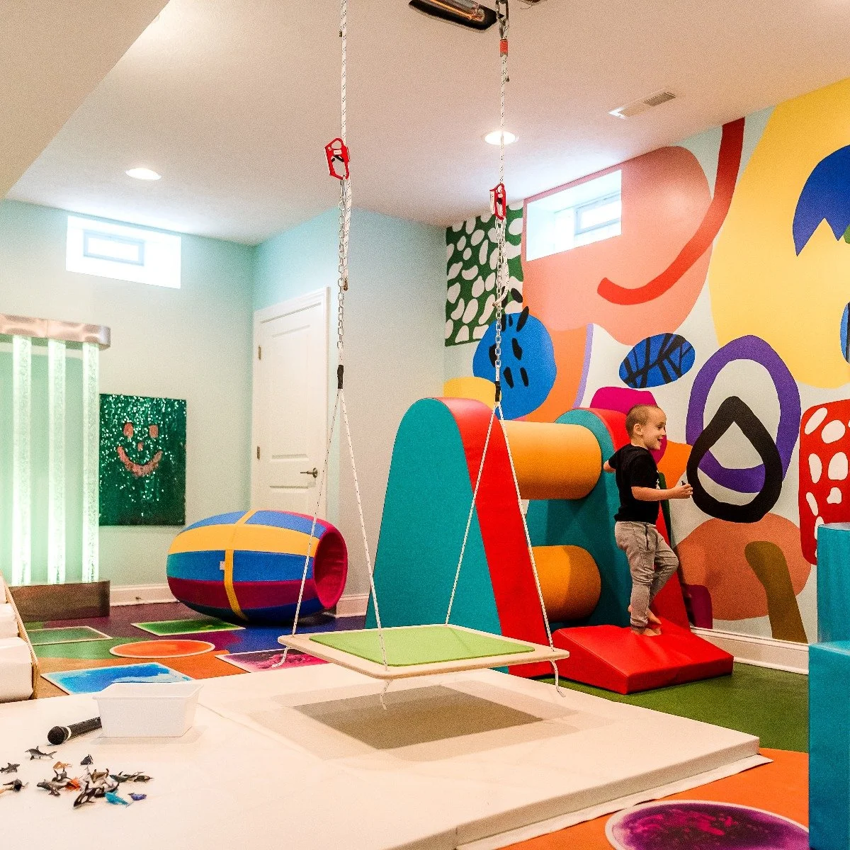 Colorful indoor children's playroom with climbing structures, a swing, and vibrant wall art, featuring a young boy playing.