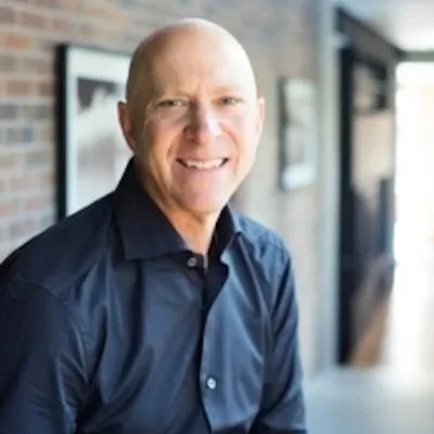 A photo of Dihedral Co-Founder Jeffrey Katz, smiling, wearing a dark blue shirt, sitting in a well-lit indoor space with a brick wall and framed pictures in the background.