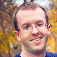 A smiling man with glasses and short dark hair outdoors, with autumn trees in the background.