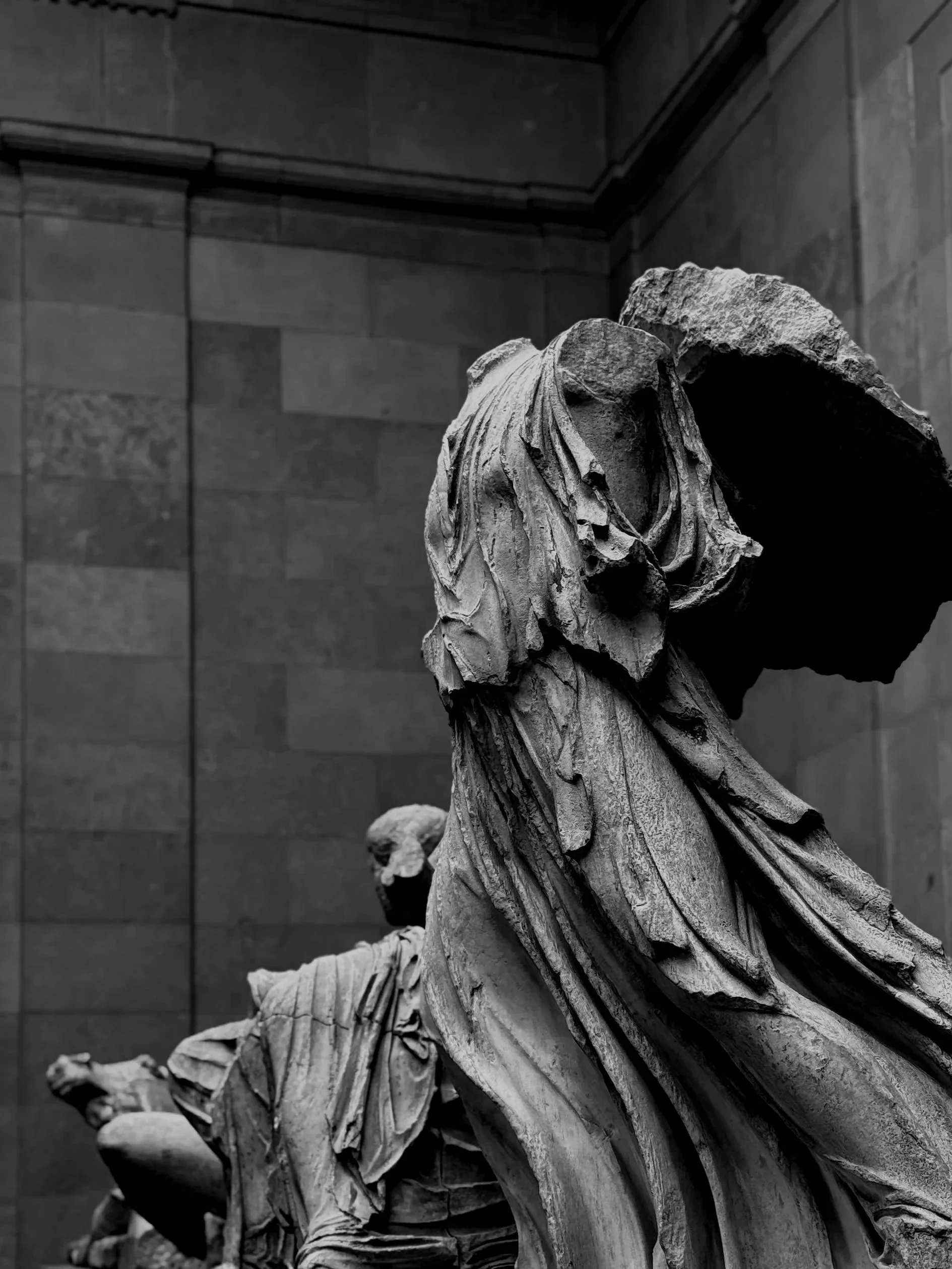 Black and white photograph of classical statue fragments in a museum, showing detailed and weathered sculpted figures with expressive drapery and broken heads.