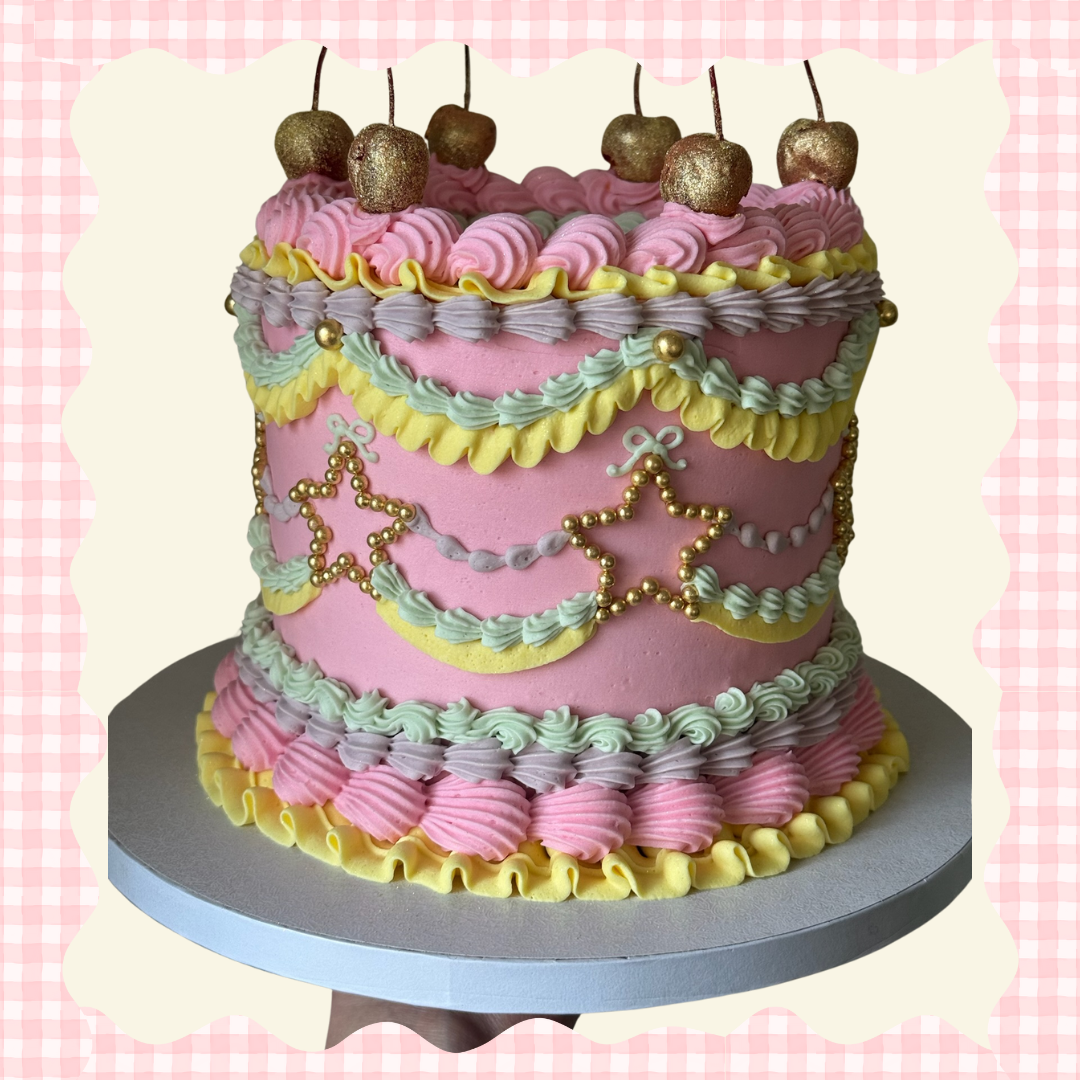 A decorated birthday cake with pink, yellow, green, and purple icing, topped with gold-colored cherries and gold beads, featuring star-shaped decorations and pastel-colored piped borders, placed on a silver cake board.
