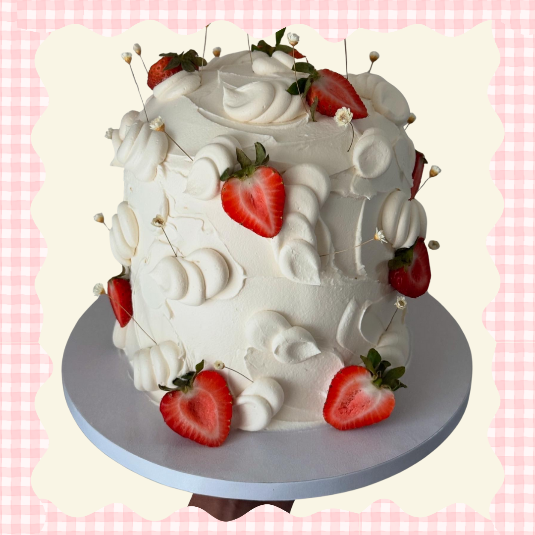 White frosted cake with strawberries and small white flowers on top, placed on a gray cake board against a pink gingham background.