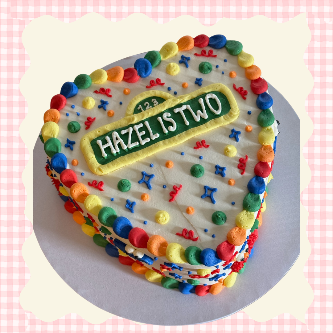 Heart-shaped birthday cake decorated with colorful frosting dots and sprinkles, with a green and yellow sign reading "Hazel is two" on top.