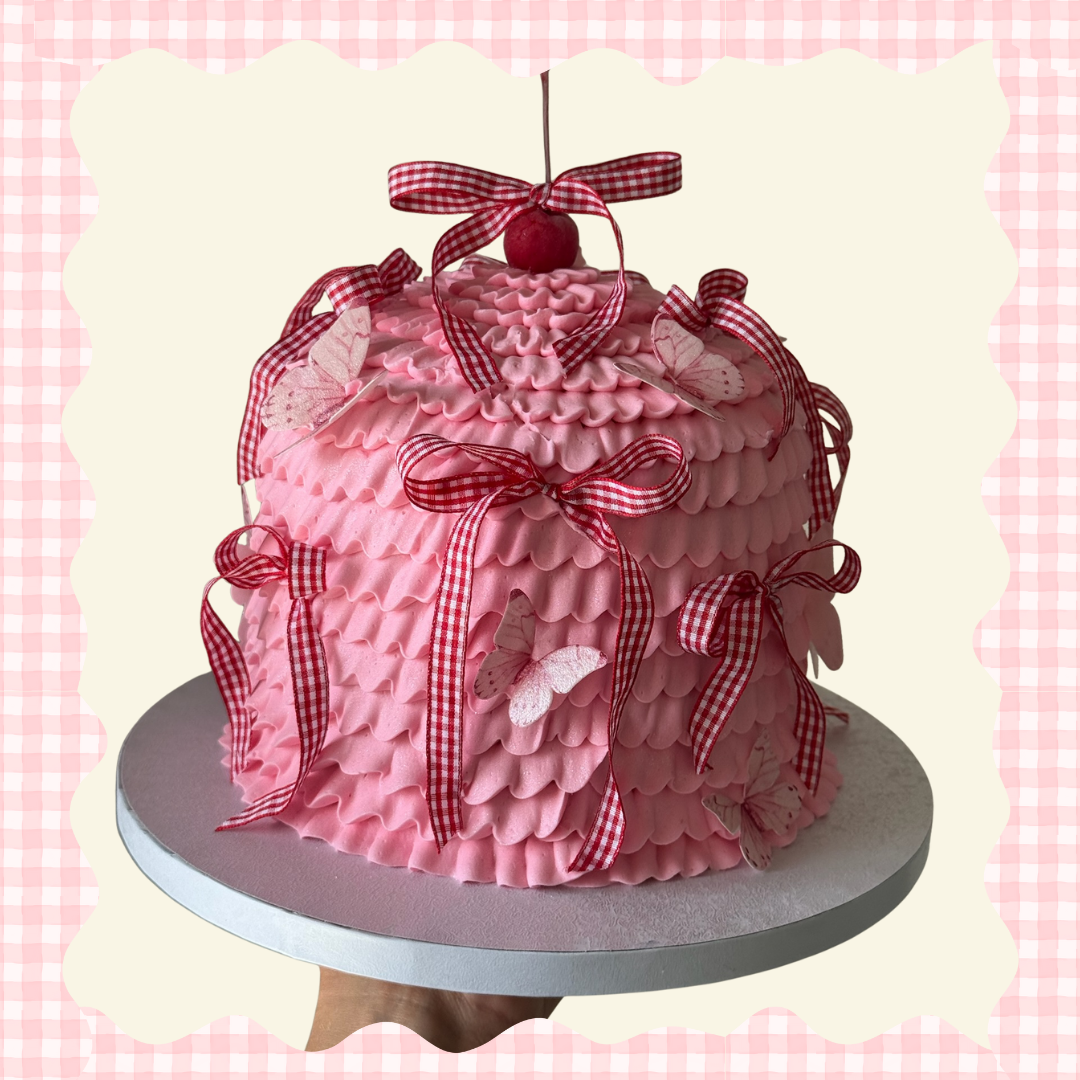Pink layered cake decorated with red and white checkered ribbons, butterfly and cherry on top, and pink icing accents, on a silver cake board.