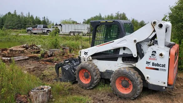 Peninsula Junk Removal Bobcat skidsteer clearing debris with dump trailer and truck in the background on the Bruce Peninsula