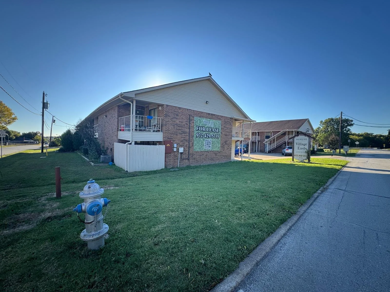 A brick apartment building with a sign indicating it is available for lease, surrounded by green lawn and a fire hydrant in the foreground, with a blue sky overhead.