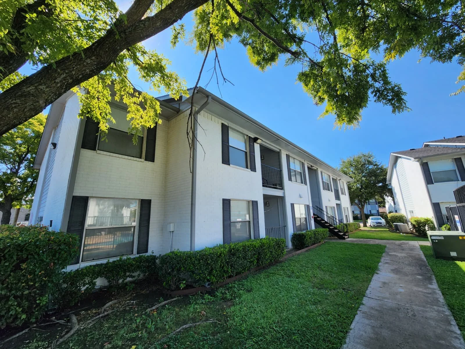 A white multi-story apartment building with black window shutters, surrounded by green shrubs and trees, under a clear blue sky.