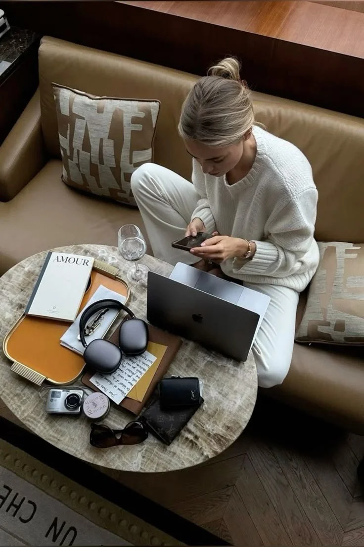 A woman sitting on a beige sofa at a round wooden table with a laptop, smartphone, glasses, headphones, camera, and various papers, in a hotel room or lounge setting.