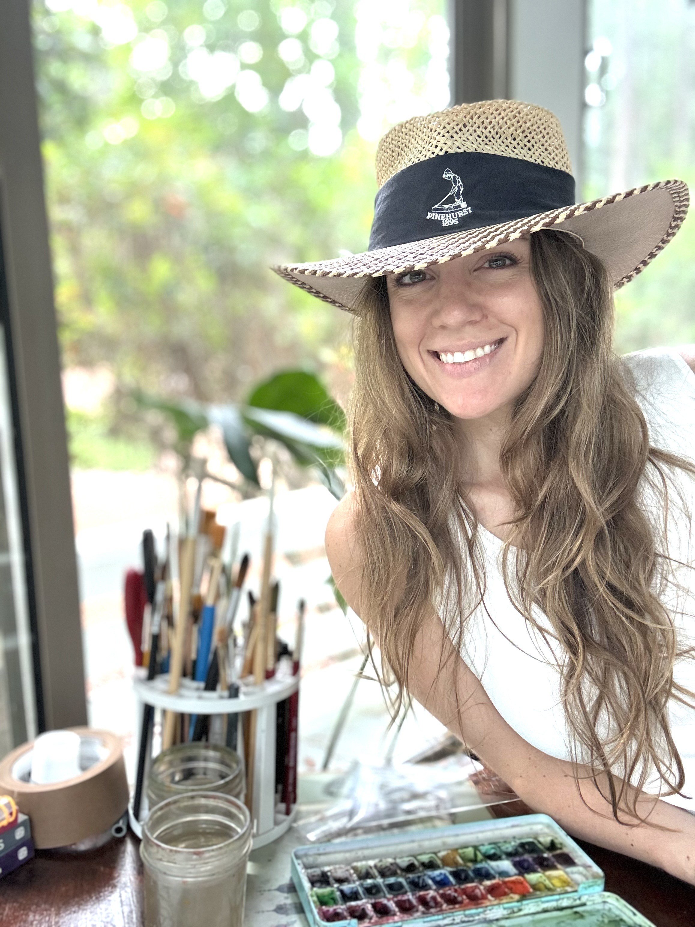 A woman smiling wearing a straw hat with a black band, sitting at a table with watercolor paints and brushes, indoors with a window showing greenery outside.