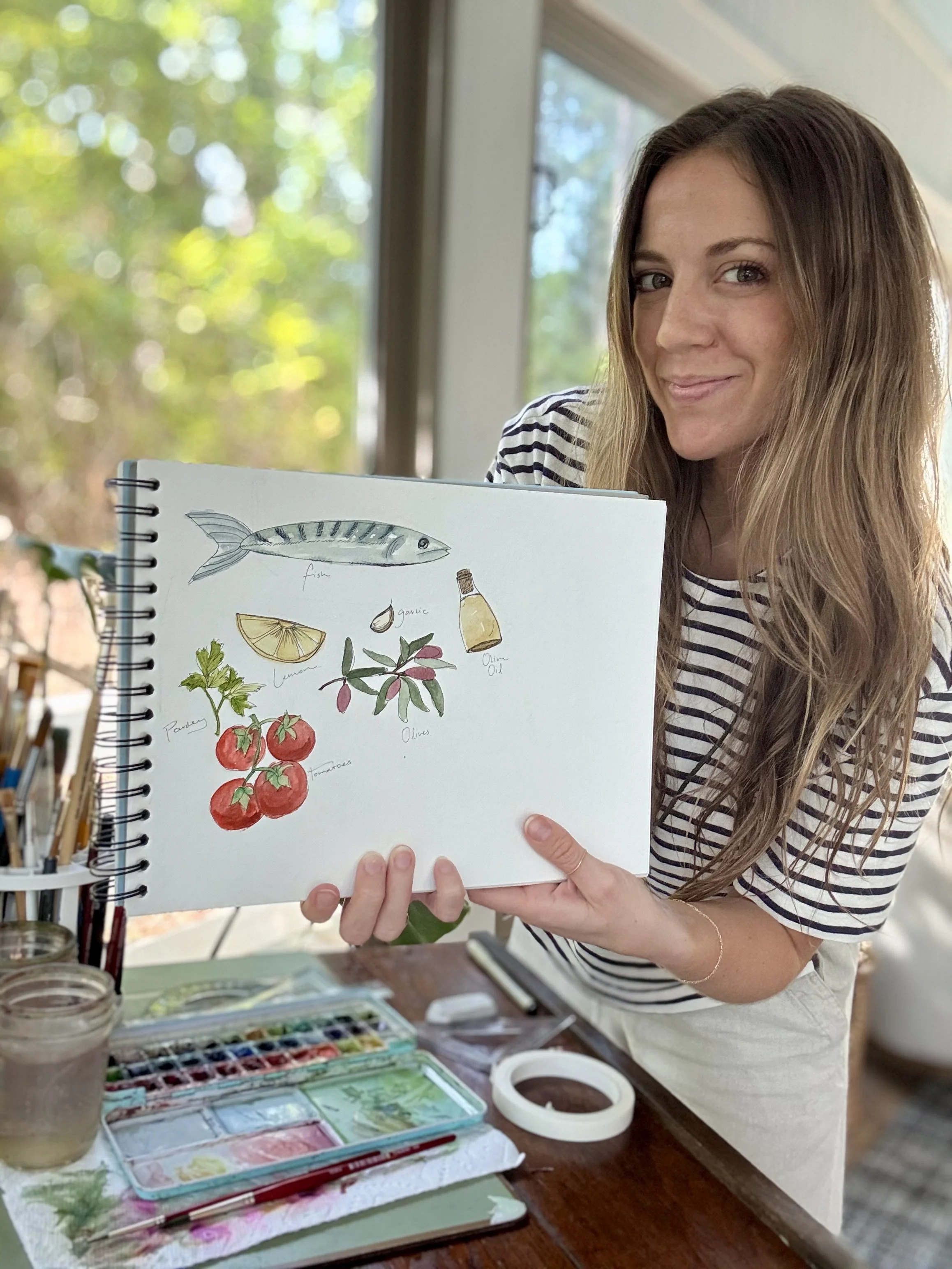 A woman with long wavy hair holding a sketchbook with watercolor illustrations of fish, lemon, garlic, olive oil, parsley, tomatoes, and olives, standing indoors near a window with a garden view.