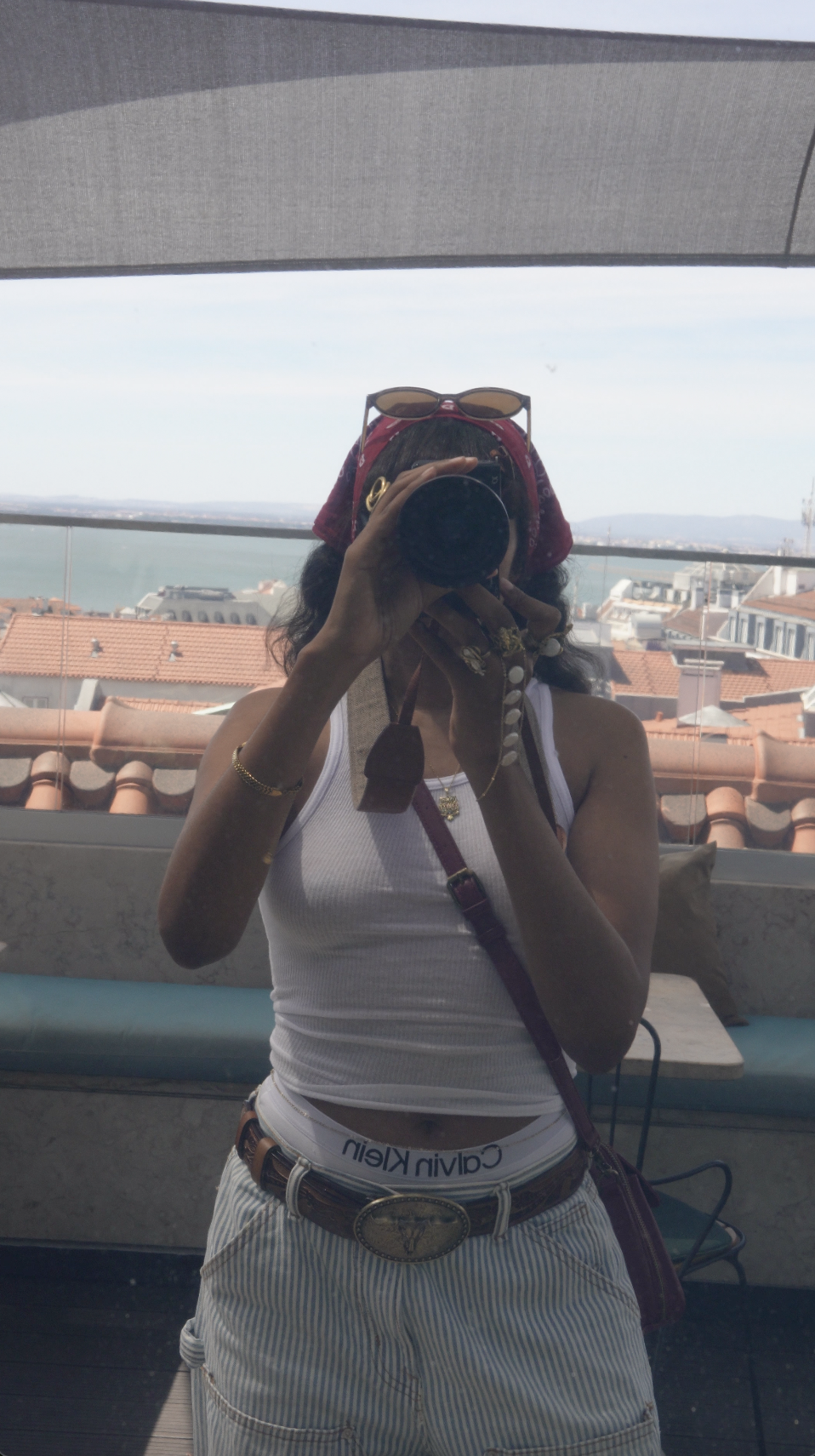 A woman taking her photo in a mirror, wearing a white tank top, striped shorts, a belt with a large decorative buckle, a red bandana, and sunglasses on her head, with a scenic cityscape and water in the background.