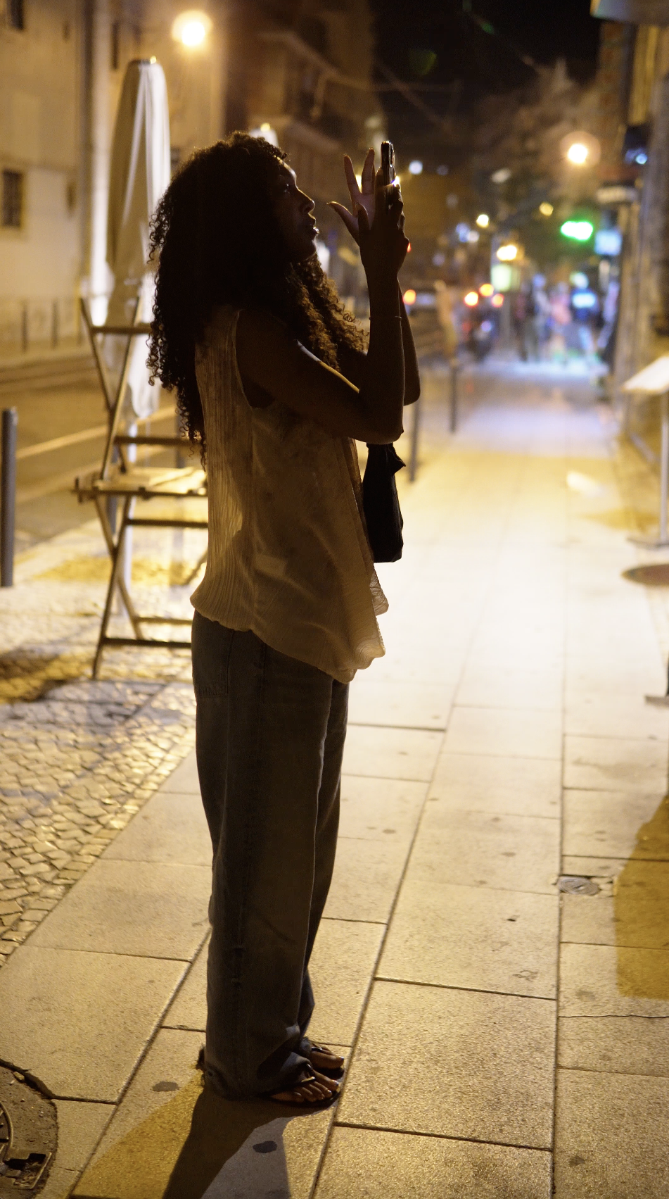 A woman standing on a city sidewalk at night, silhouetted against streetlights, with her hands raised near her face.