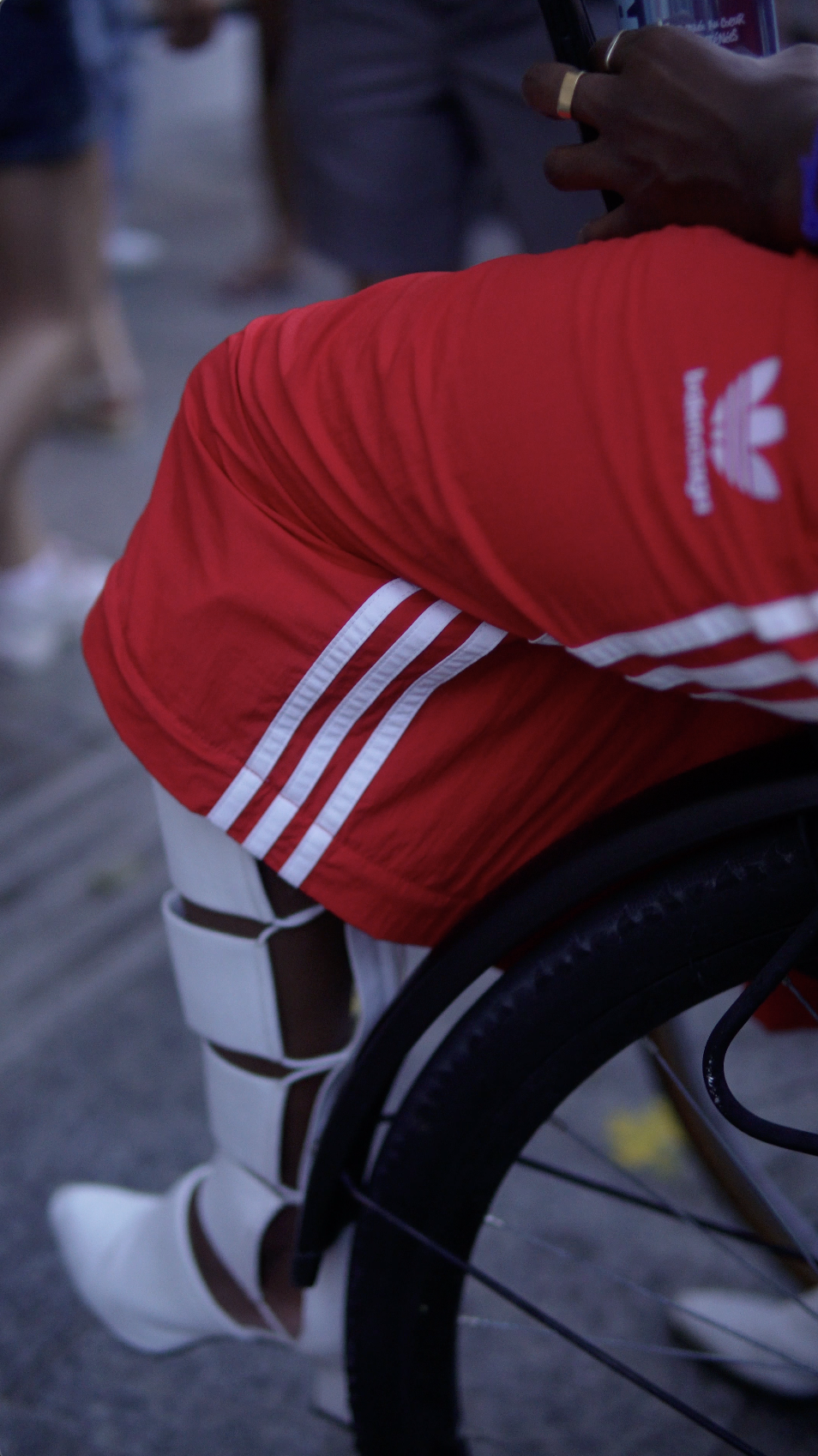 Close-up of a person in a wheelchair wearing white knee-high gladiator sandals, red Adidas sports shorts, and holding a phone in their hand, with a blurred background of other people.