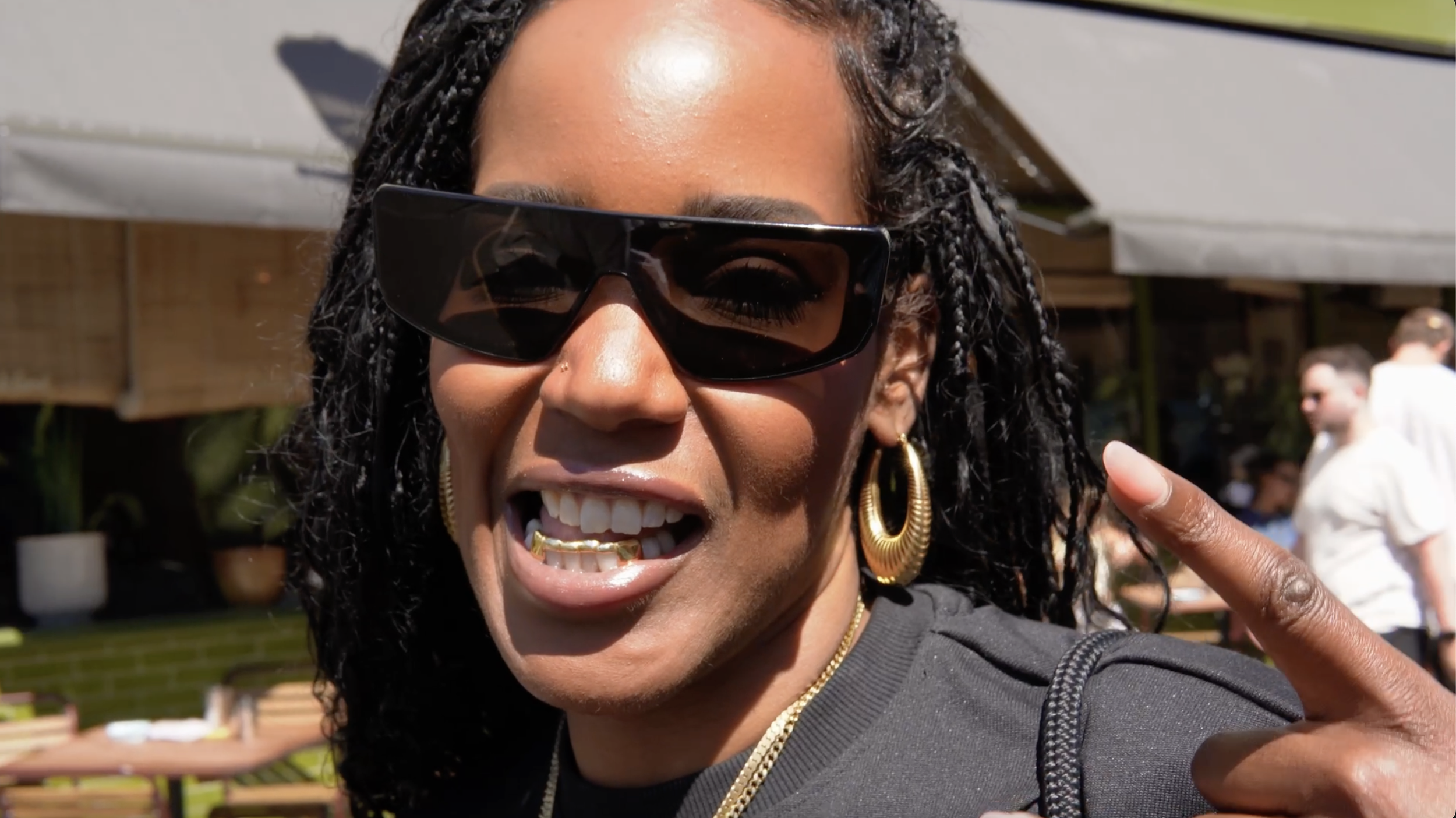 Close-up of a smiling woman with curly hair wearing large sunglasses and gold jewelry, pointing a finger in the air outdoors on a sunny day, with people and trees in the background.