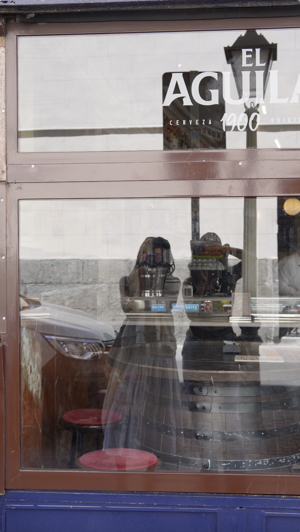 Photo of a restaurant window with signage reading "El Aguila 1900" and "Cerveza." Inside, a person is reflected, wearing a striped dress and standing near a bar or counter with red stools and bottles on shelves.