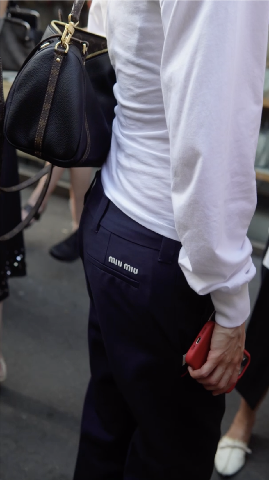 Close-up of a person wearing black Miu Miu pants, a white shirt, holding a red phone, and carrying a black handbag over their shoulder, at an outdoor event.