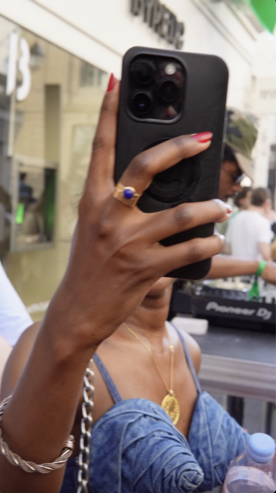Person taking a selfie at an outdoor event, holding a phone with a black case. They are wearing a gold necklace with a large pendant, a gold bracelet, a blue denim jacket, and a ring with a blue stone. In the background, there are other people, one w