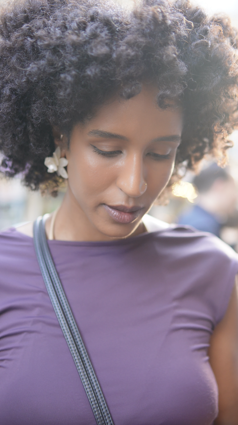 A young woman with curly hair, earrings, and a nose ring wearing a purple shirt and a shoulder strap, looking down with a serene expression."}