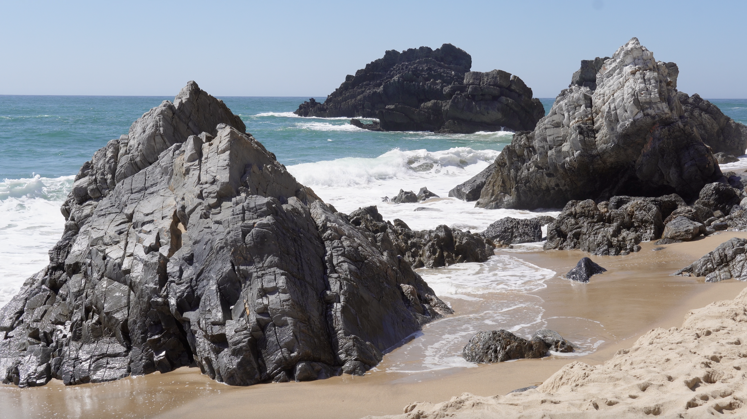 Rock formations on a sandy beach with ocean waves crashing against them under a clear blue sky.