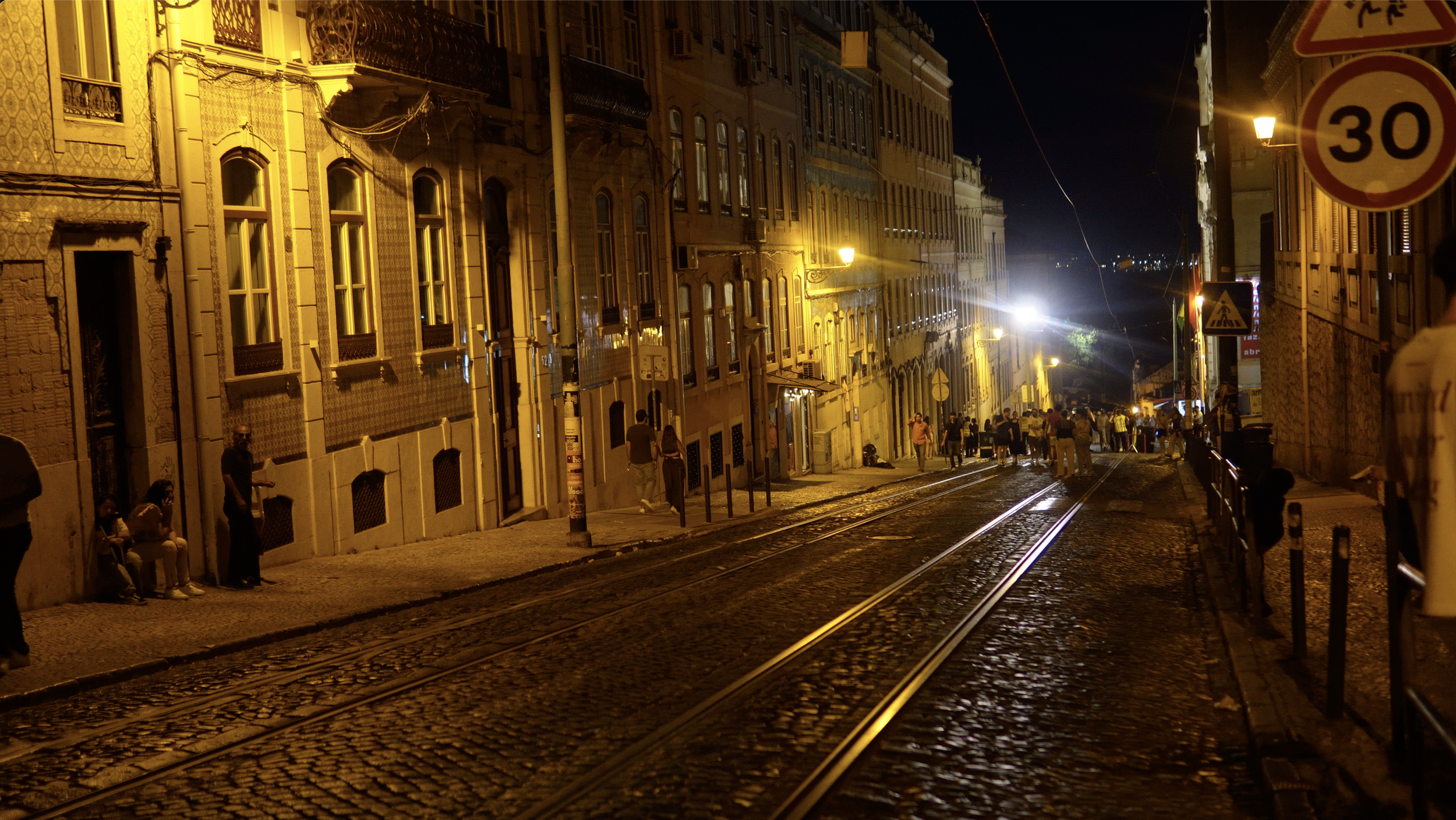 Night view of a street with tram tracks, illuminated by yellow streetlights, lined with old buildings, with people walking and sitting along the sidewalk, and traffic signs including a 30 km/h speed limit and pedestrian crossing signs.