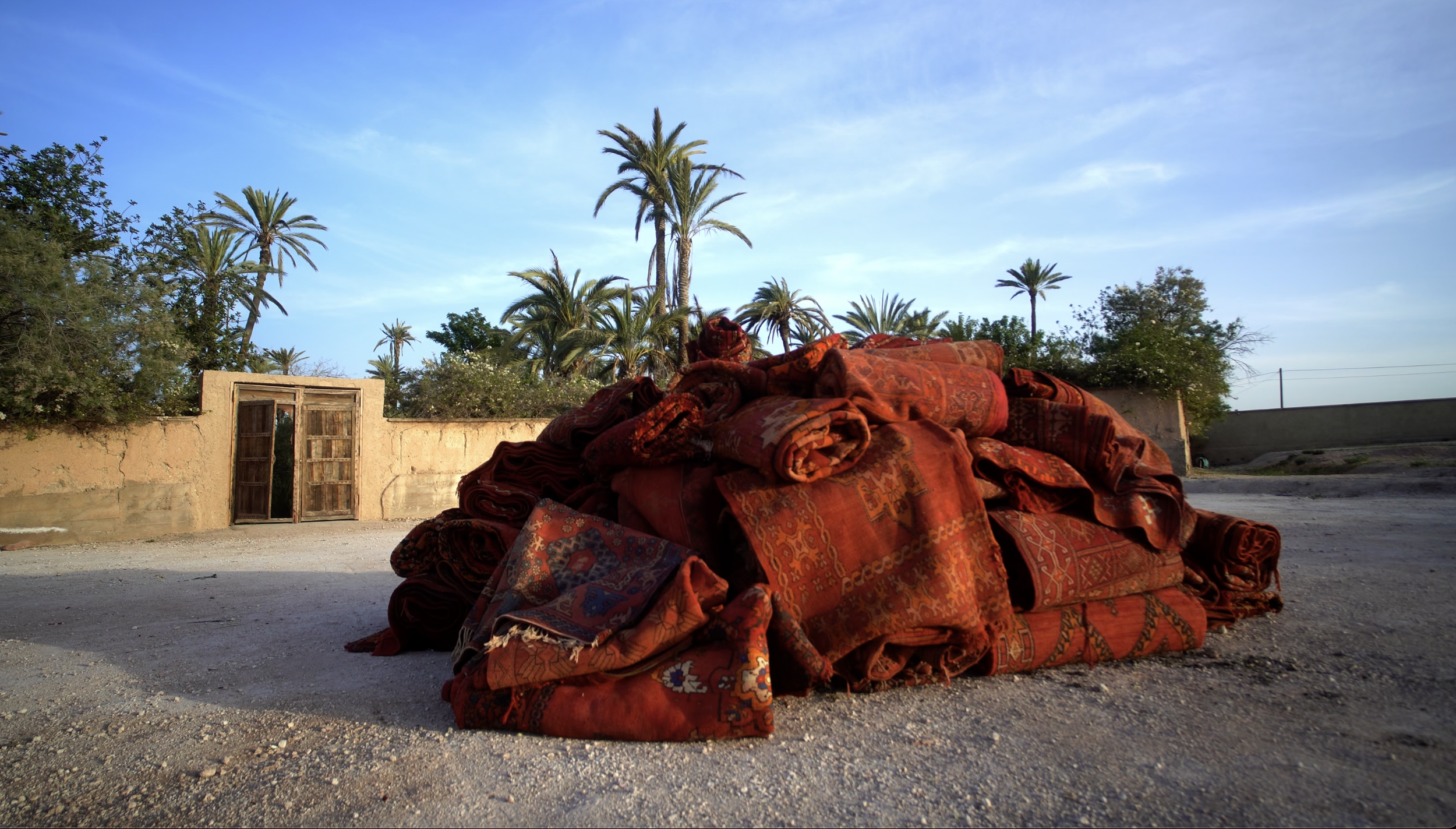 Large pile of red and orange patterned rolled-up carpets on dirt ground, with palm trees and a concrete wall in the background under a blue sky.