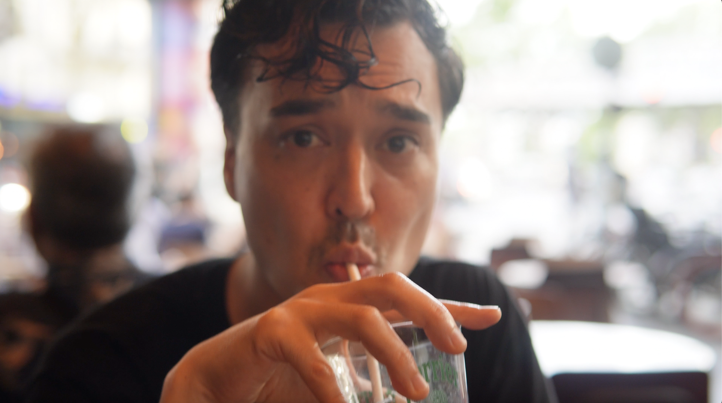 A young man with dark, curly hair drinking a beverage through a straw in a cafe or restaurant setting.