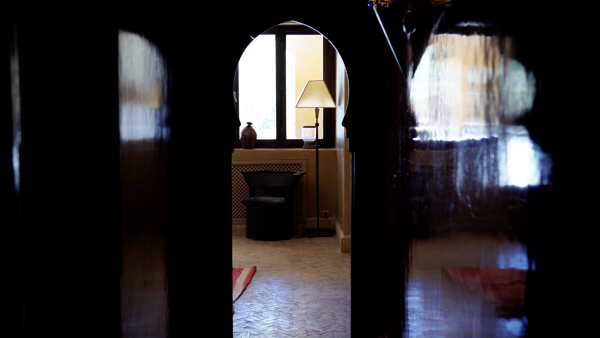 View through a dark doorway into a room with a window, a lampshade, a chair, and decorative items on the windowsill.