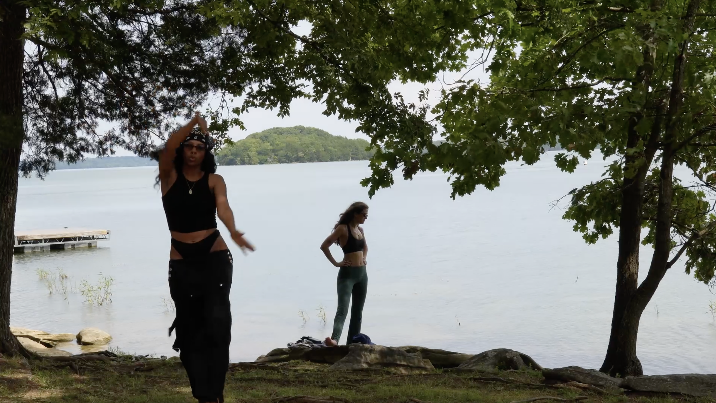 Two women near a lakeshore under trees, one in black attire with sunglasses and headband, another in yoga clothes, with a dock and lake in the background.