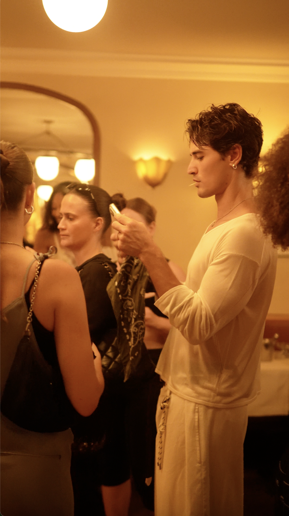 A young man with short, curly hair and earrings is looking at his phone while standing in a dimly lit, warm-colored room with several people around him. He is wearing a light-colored shirt and pants. The room has an arched doorway, hanging lights, an