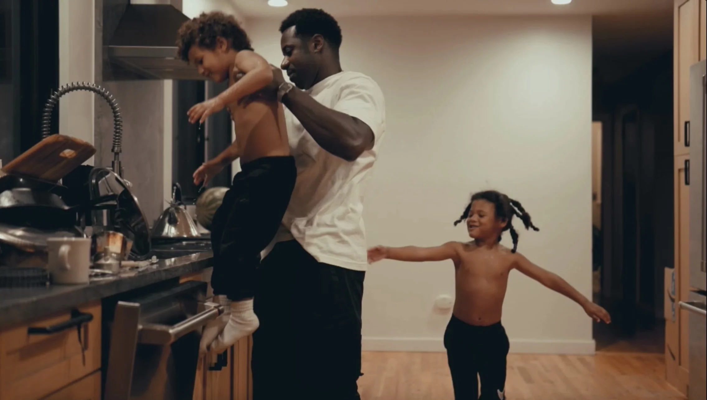 A man and two children in a kitchen, the man is helping one of the children wash his hair at the sink, while the other child plays behind them.