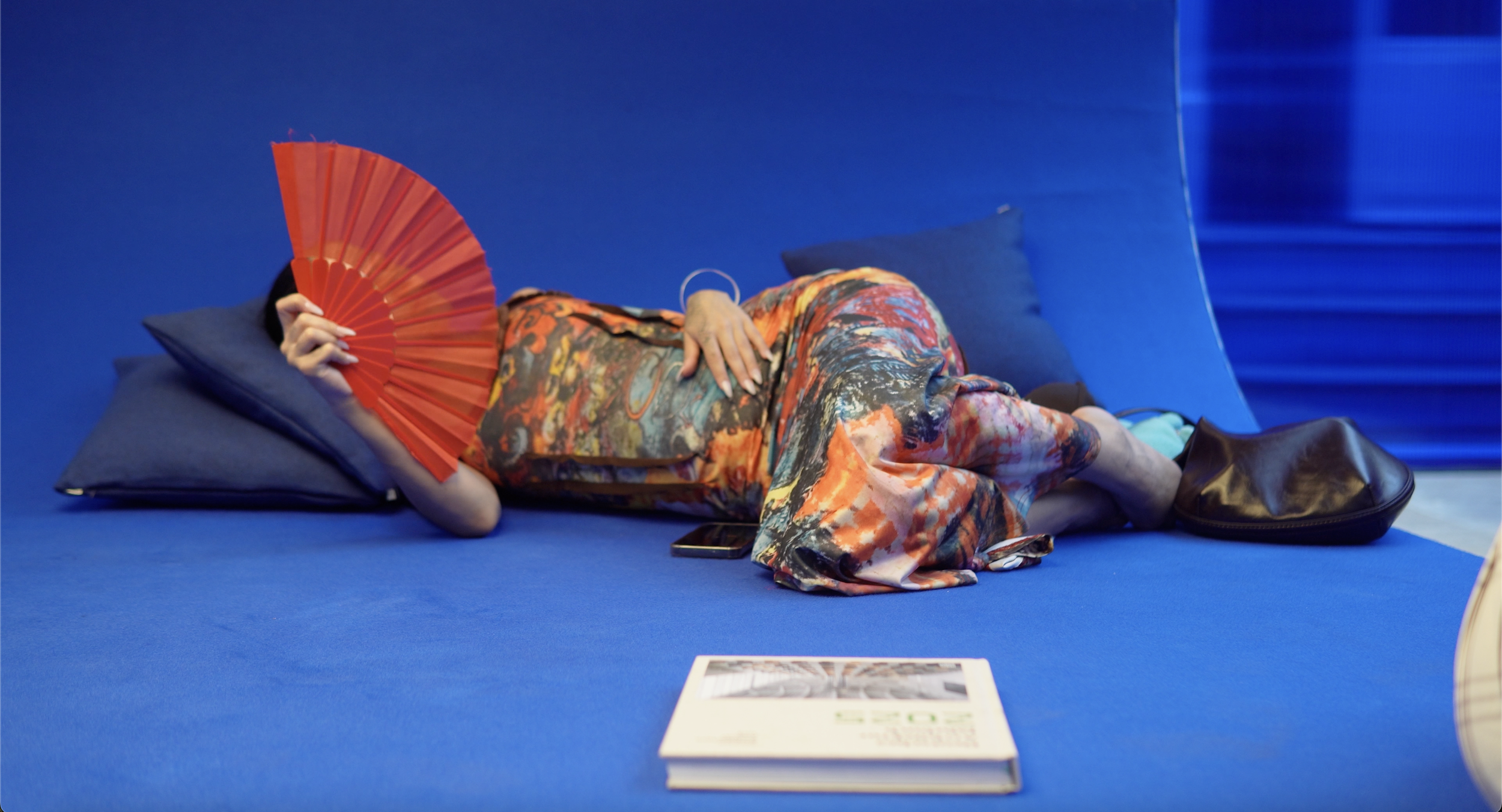 Person lying on a blue floor with pillows, holding a red fan to their face, wearing a colorful, abstract-patterned dress, with a phone nearby and a magazine or booklet in front.
