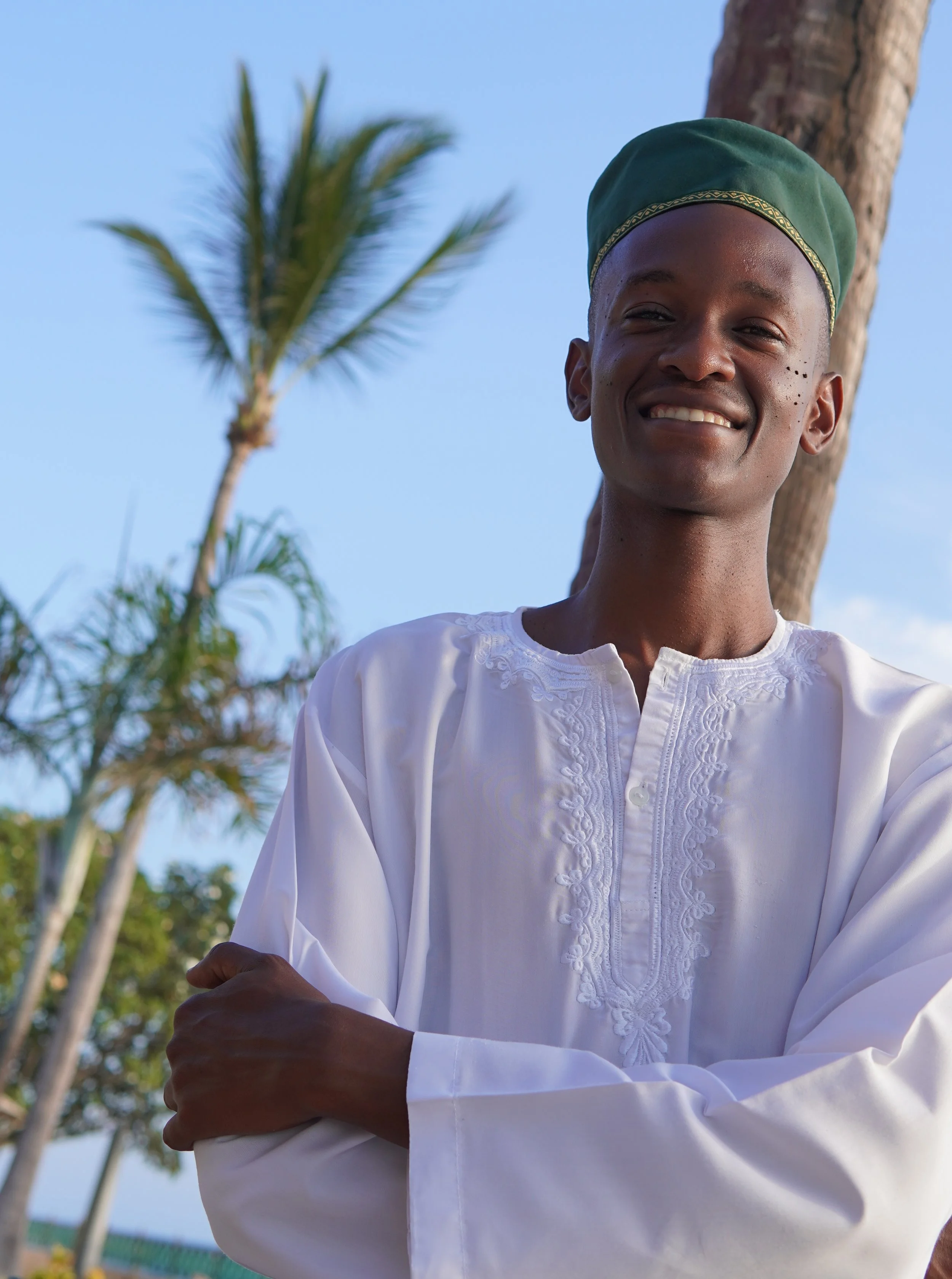 A smiling person wearing a white traditional garment and a green hat, standing outdoors with palm trees and a clear blue sky in the background.