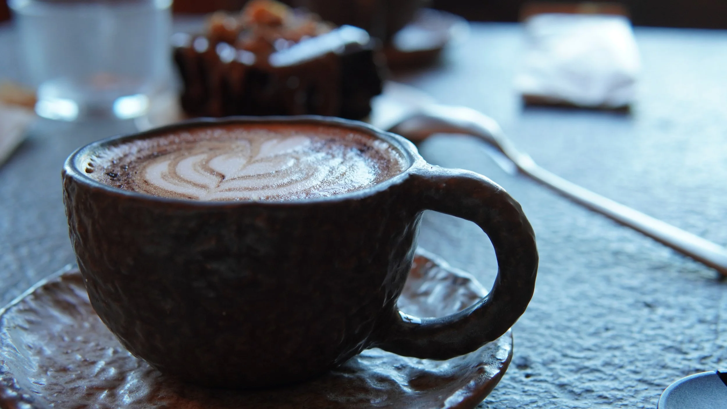 A ceramic cup filled with coffee topped with foam and latte art, placed on a saucer on a table with blurred background.