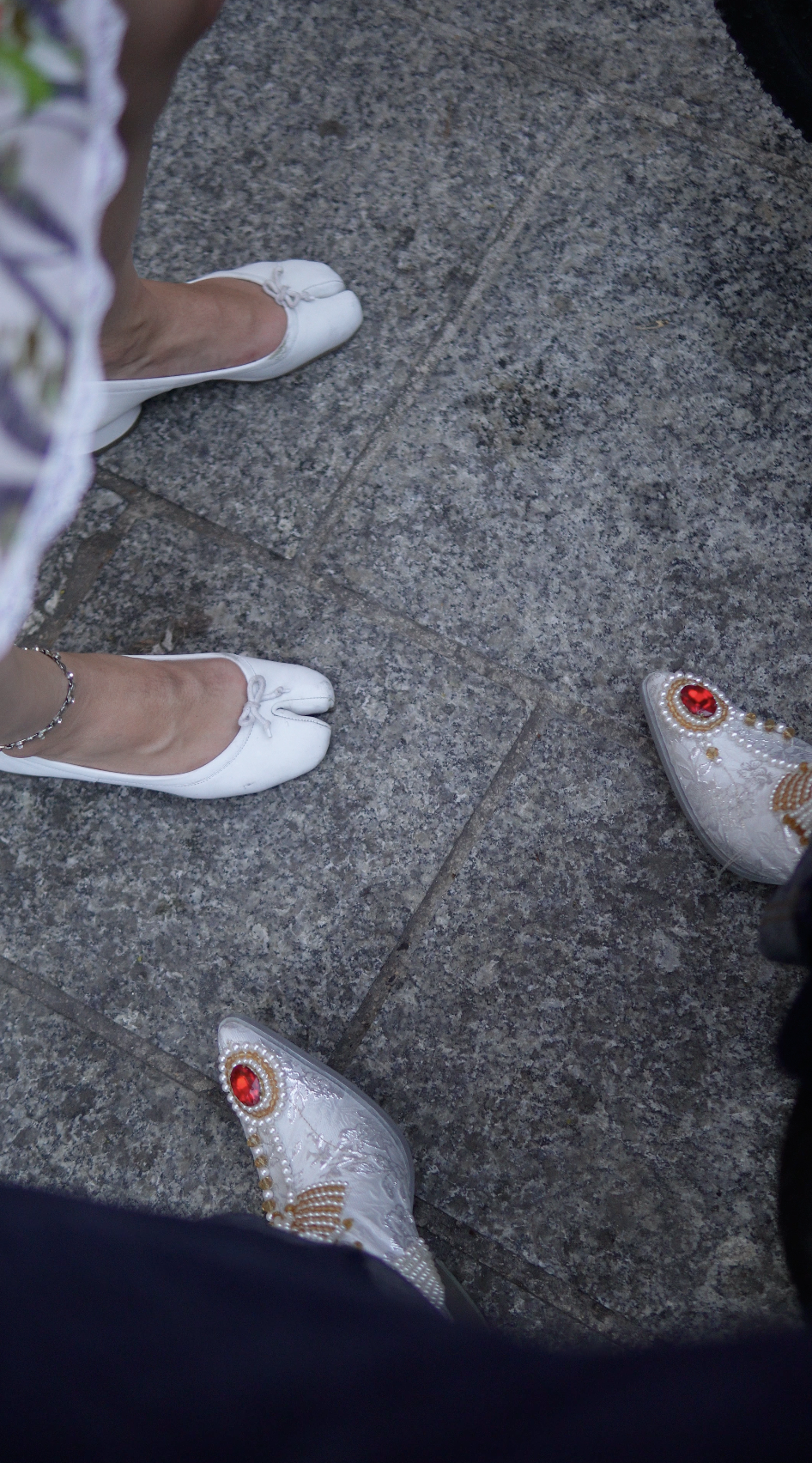 View of two women wearing white ballet flats with bows, one with a silver ankle bracelet, standing on a gray stone pavement. A third person's ornate white shoes with red gemstones and gold accents are partially visible.