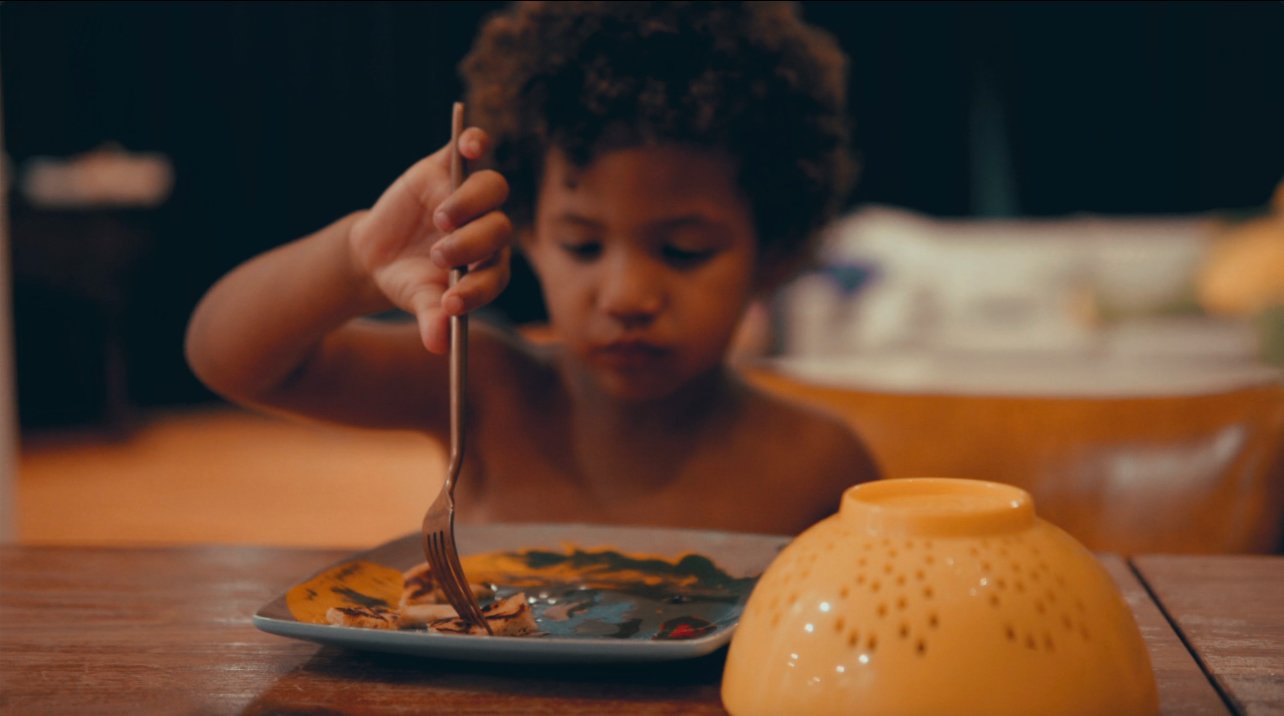 A young child with curly hair is sitting at a wooden table, using a fork to eat food from a plate.