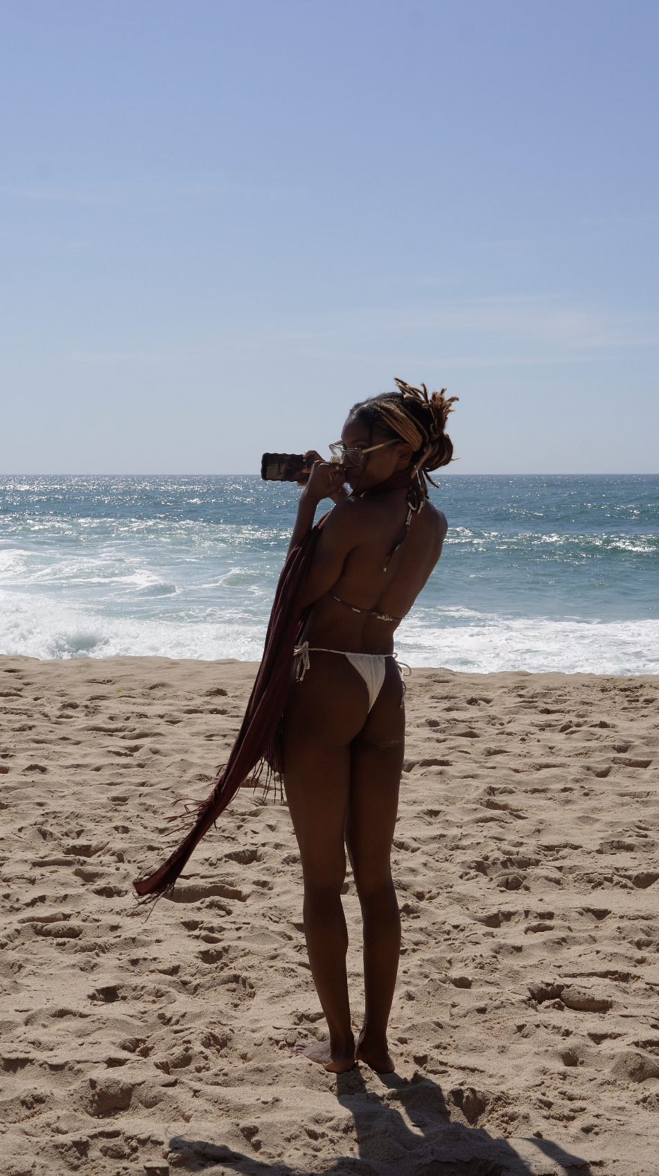 A woman with dreadlocks and glasses standing on the sandy beach, holding a camera, with the ocean and blue sky in the background.