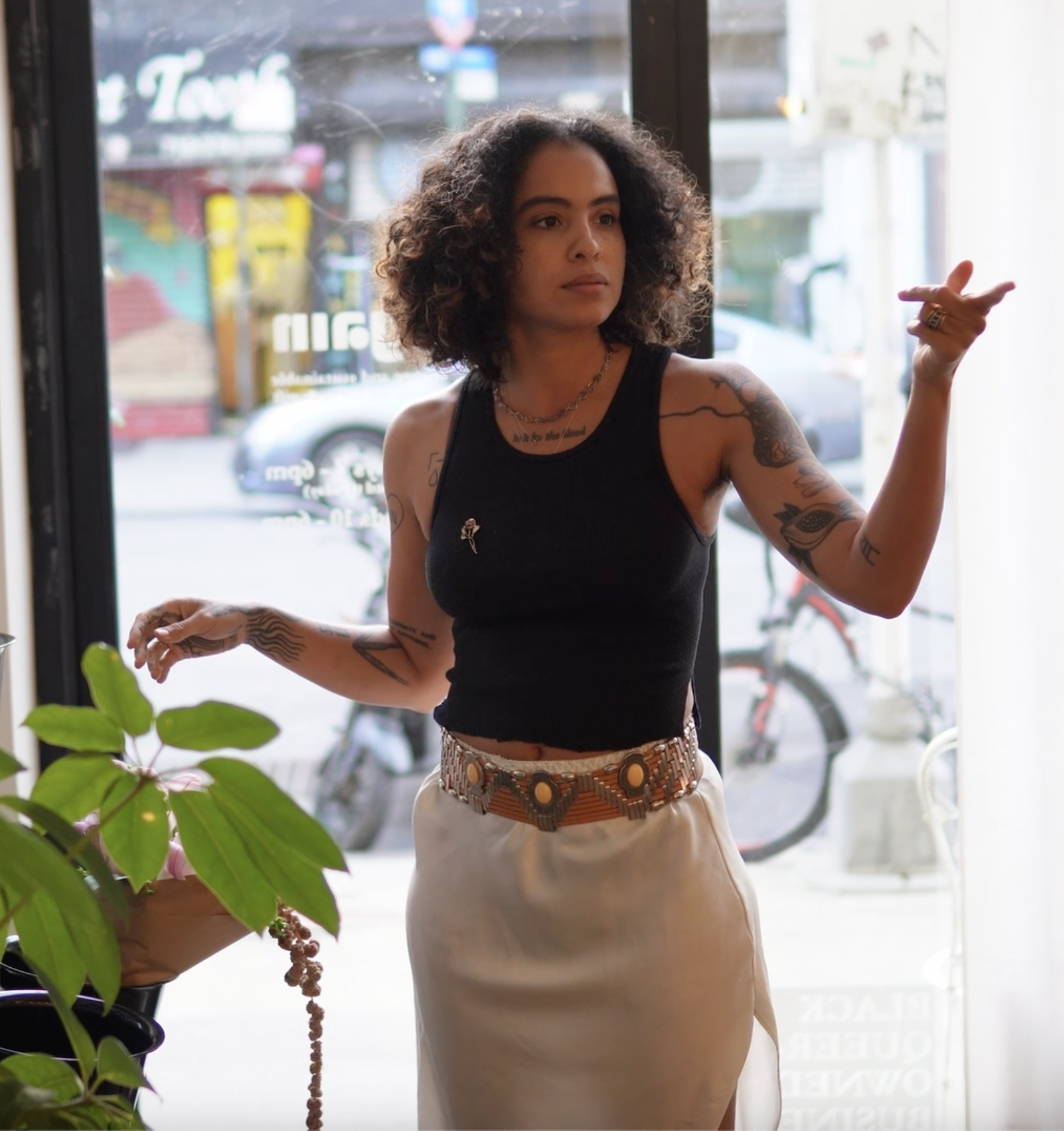 A woman with curly hair, wearing a black sleeveless top and beige skirt, standing indoors near a glass door, gesturing with her right hand, and surrounded by plants.