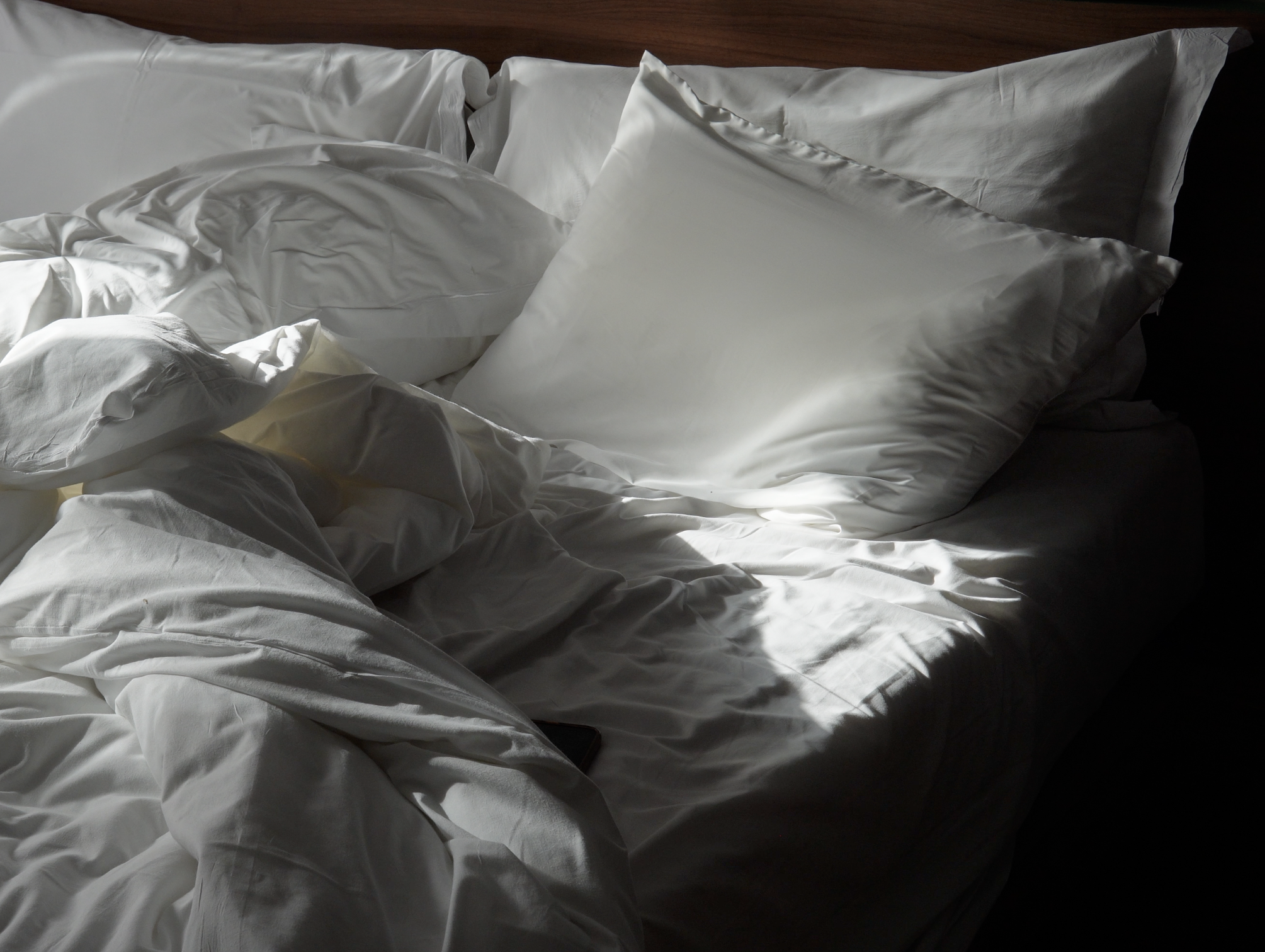 Unmade white bed with rumpled sheets and pillows, lit by natural light with shadows.