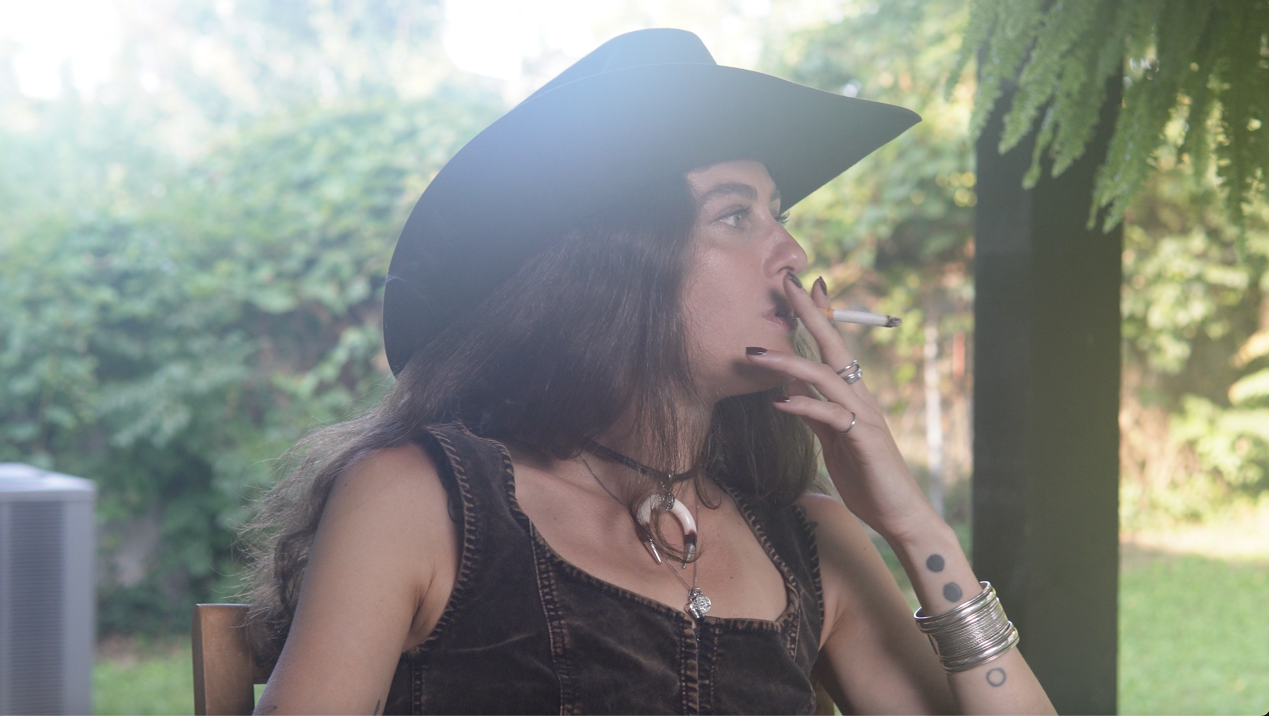 A woman with long dark hair wearing a witch's hat, smoking a cigarette, and wearing layered necklaces and bracelets, sitting outdoors with green trees in the background.