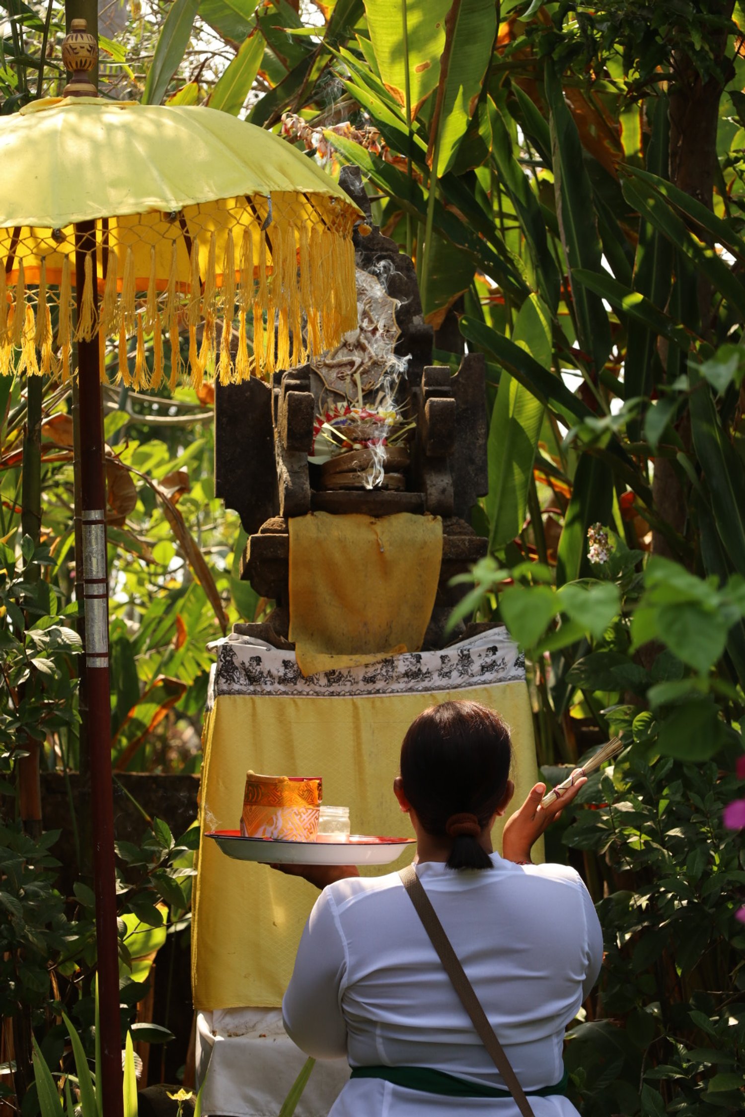 A person performing a traditional ritual or offering in a lush, green outdoor setting with various plants and trees, featuring a small shrine with smoke and religious items, a yellow parasol, and a tray with offerings.
