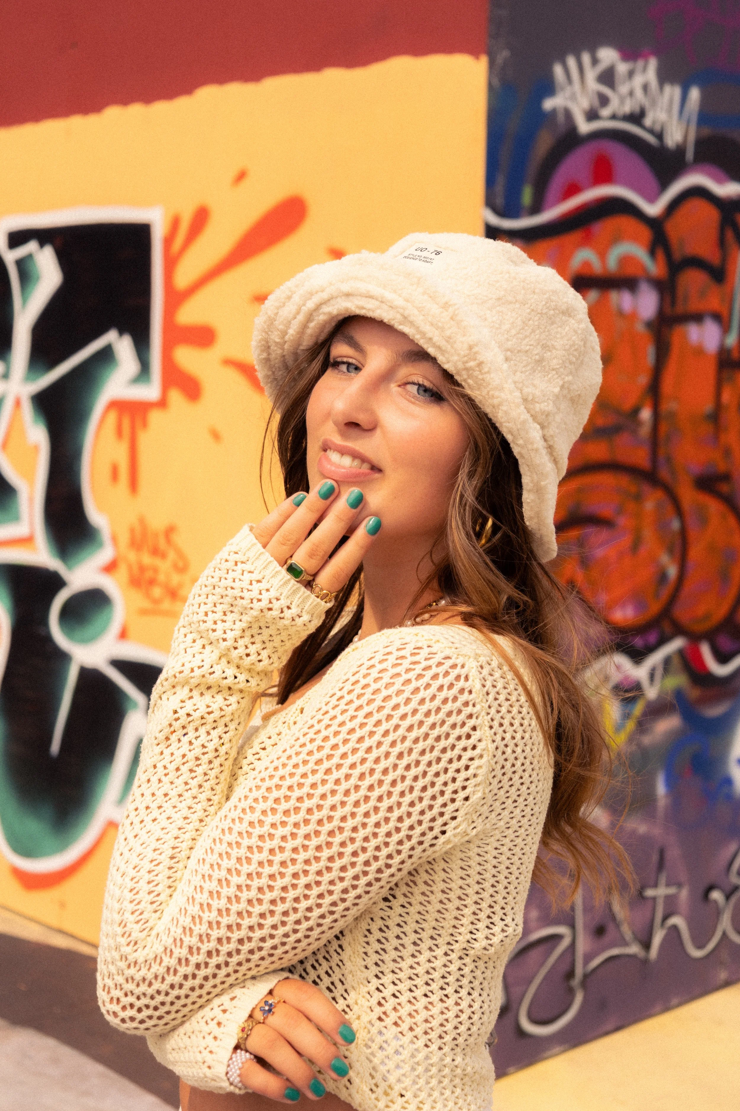Young woman posing in front of colorful graffiti art, wearing a beige knit sweater, a fluffy cream bucket hat, and turquoise nail polish.