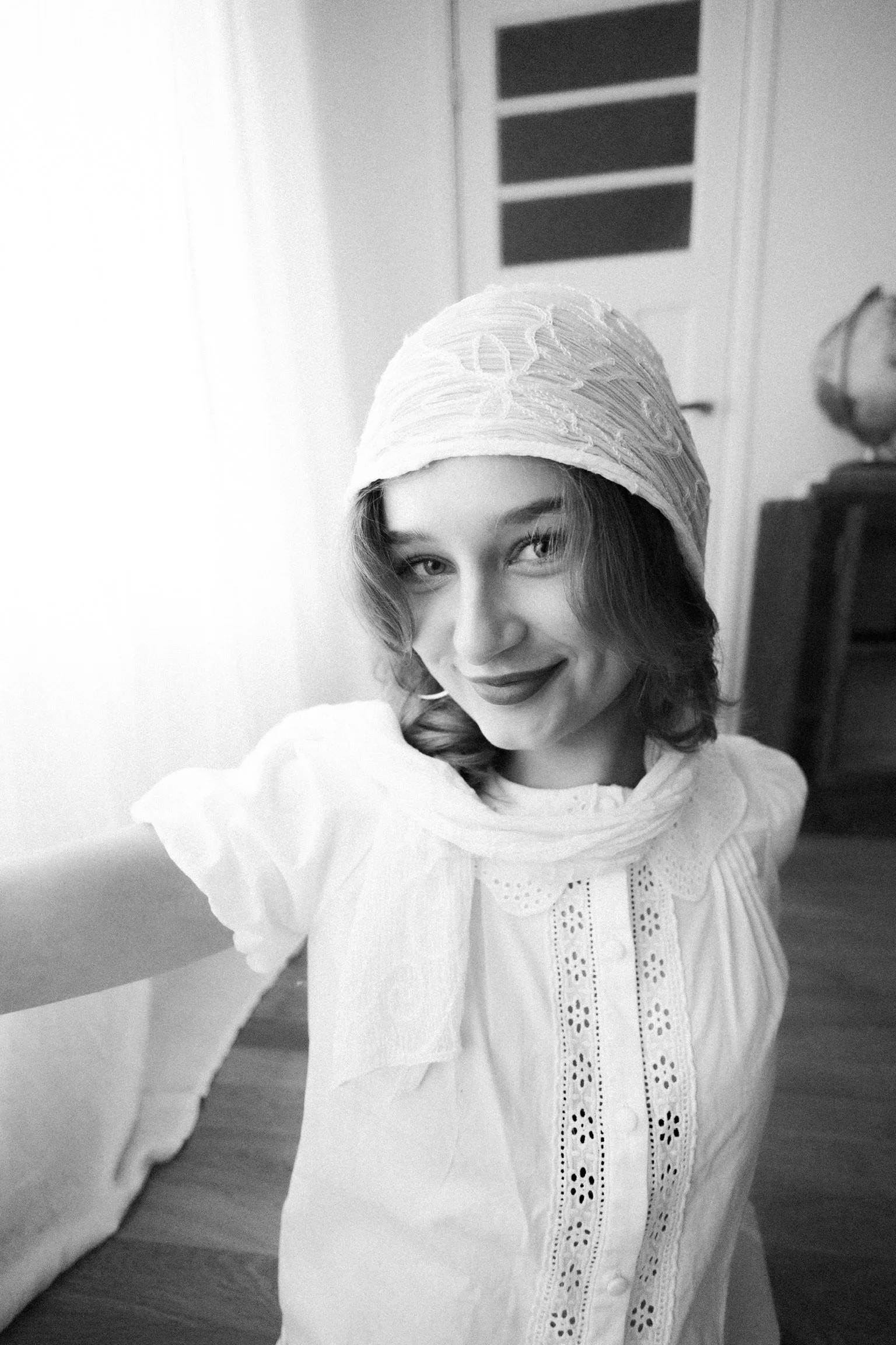 Black and white photo of a woman with shoulder-length hair wearing a headscarf and a vintage lace-collared blouse, standing in a room with a wooden floor and a dresser with a globe on top.
