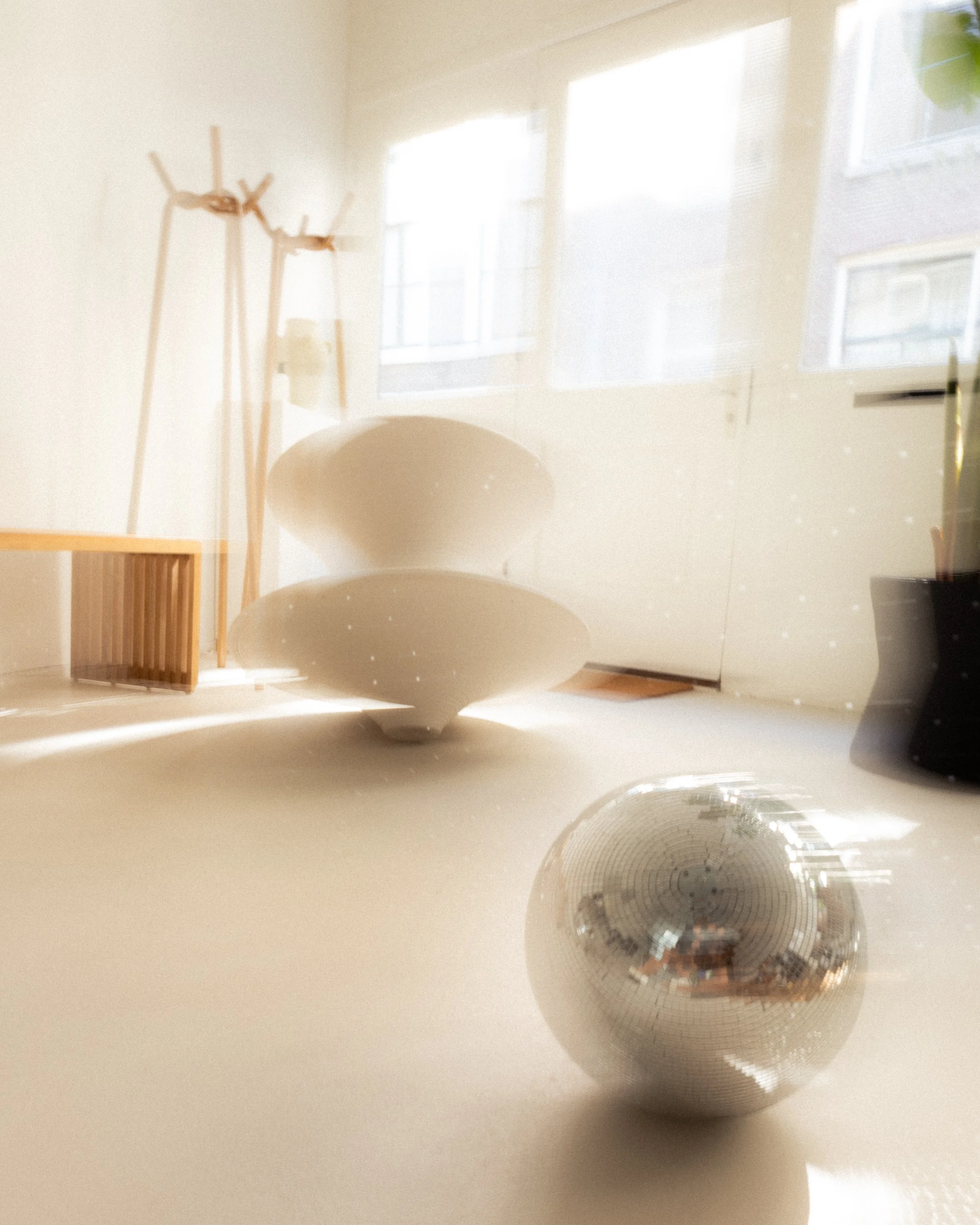 A shiny silver disco ball on the floor with sunlight reflecting off it, in a room with white walls, large windows, and minimal furniture.