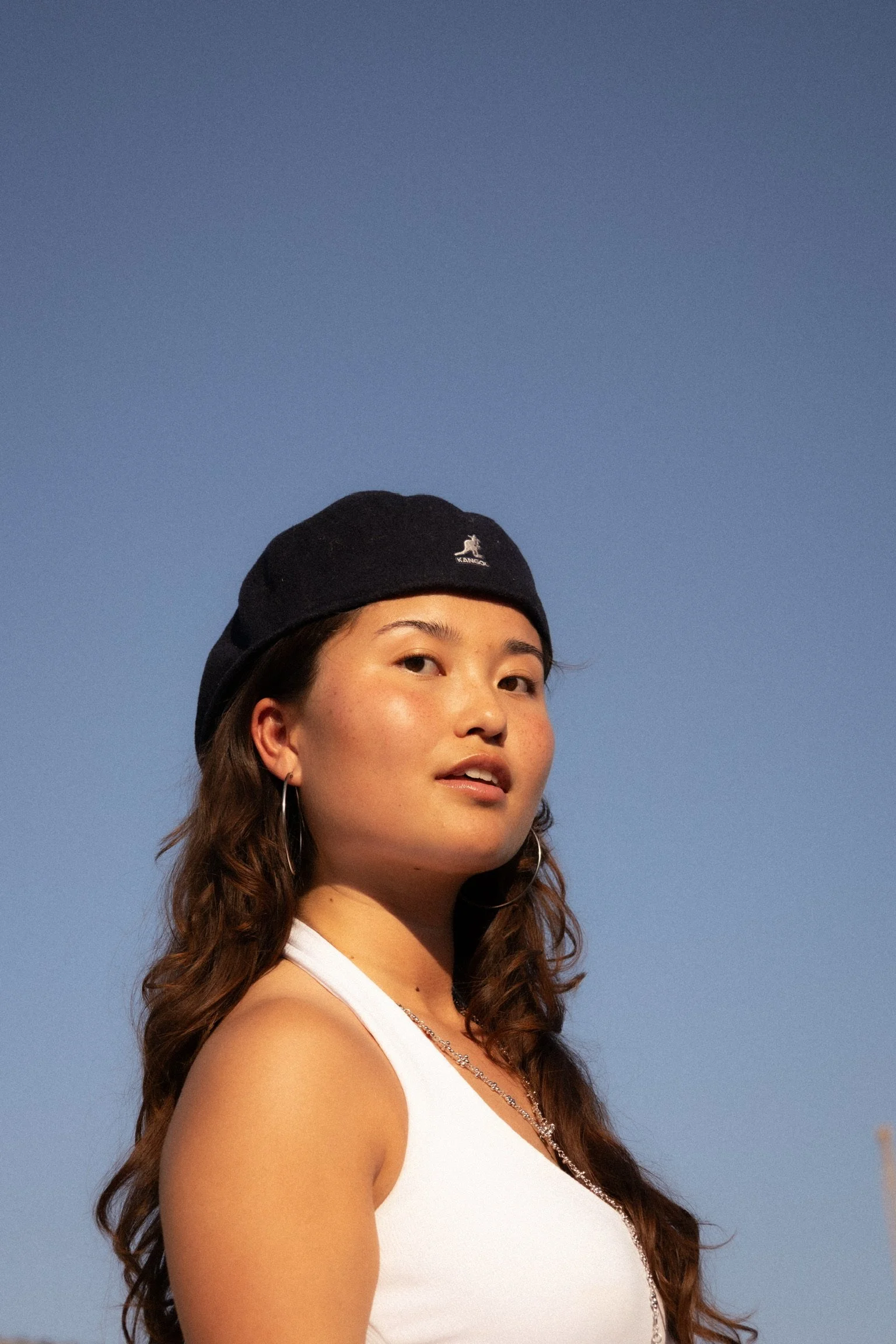 A woman with long wavy hair wearing a black beanie, hoop earrings, and a white tank top against a clear blue sky.