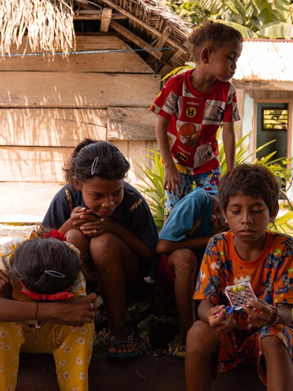 Five children sitting and standing outdoors near a wooden house, with one boy holding a small game console or electronic device.
