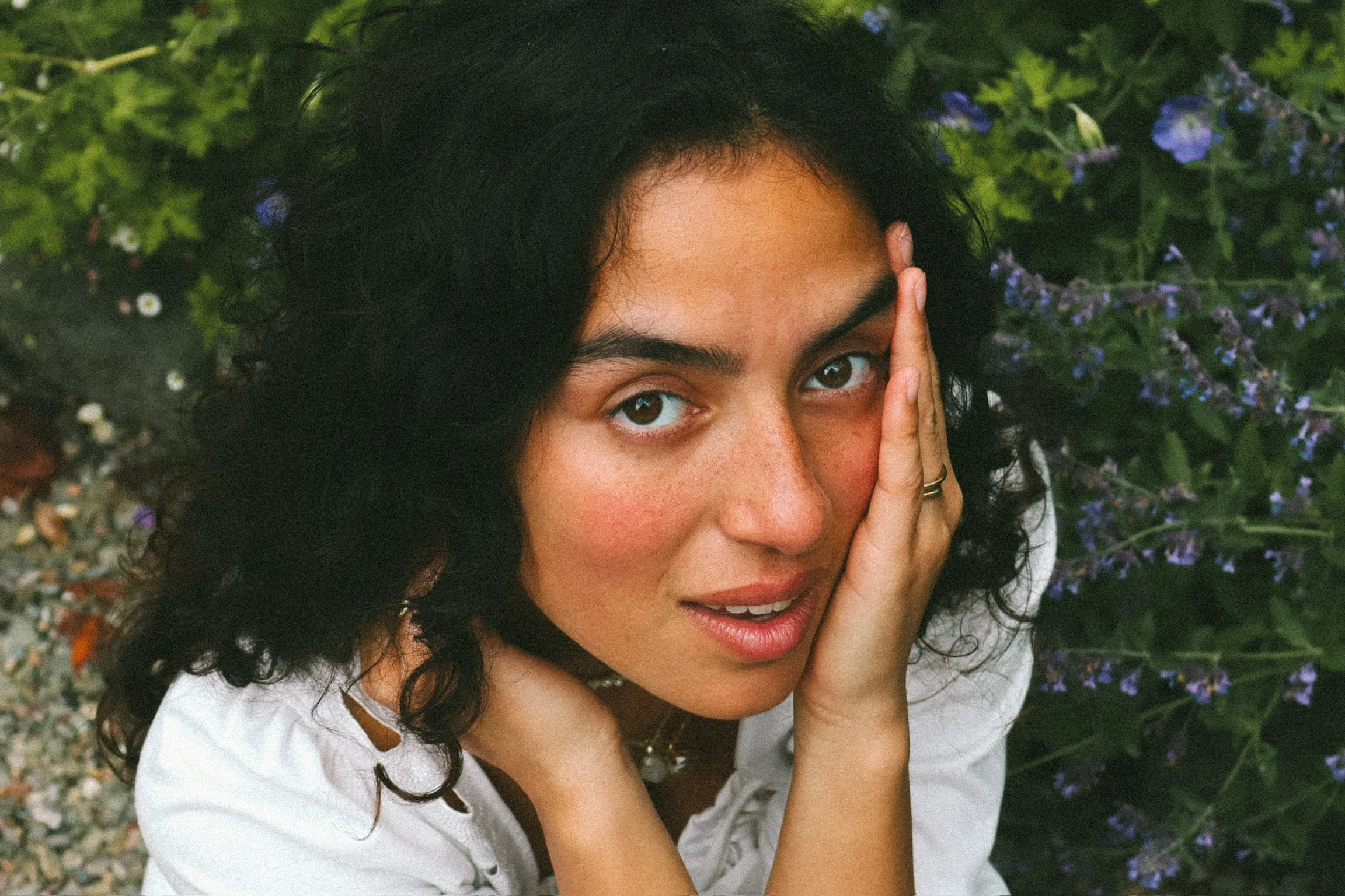 A woman with dark curly hair and freckled skin looks at the camera with one hand resting on her cheek, surrounded by green foliage and purple flowers.